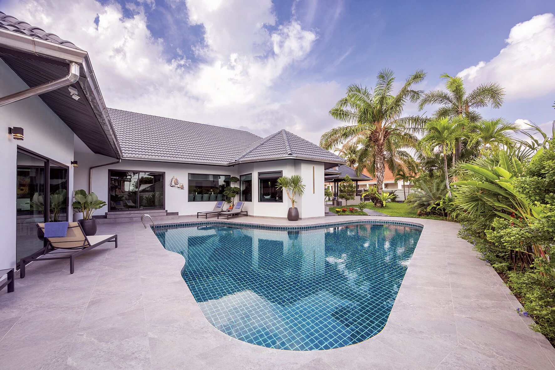 A modern house with a swimming pool, large windows, and a garden with tropical plants.