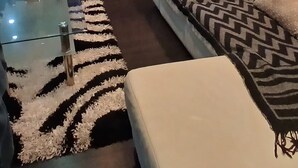 A modern living room with a large flat-screen TV on a wooden stand, a glass coffee table, a white sofa, and a patterned rug.