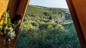 A view from a window overlooking a lush green landscape with a hill and a cliff. - Kudhva 4 Summary:<br>Per (Tintagel)