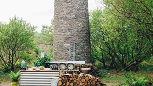 A rustic outdoor setting with a stone pillar, a wooden table with various items, and a stack of firewood. - Kudhva 4 Summary:<br>Per (Tintagel)