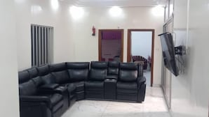 Un saló amb un sofà seccional de cuir negre, un televisor de pantalla plana muntat a la paret i una cuina visible a través d'una obertura.