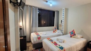 A hotel room with two beds, each with a floral bedspread and pillows, a window with curtains, a bedside table with a lamp, and a wall-mounted shelf with a decorative item. - Oasis Inka Machupicchu (Aguas Calientes)