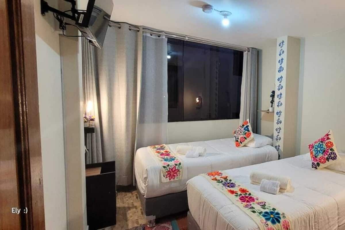 Habitación de hotel con dos camas, cada una con cubrecama y almohadas estampadas con motivos florales, una ventana con cortinas, una mesita de noche con lámpara y un estante de pared con un objeto decorativo.