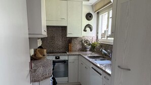 Une cuisine étroite avec des meubles blancs, un plan de travail en granit, un four encastré et une fenêtre équipée de stores.