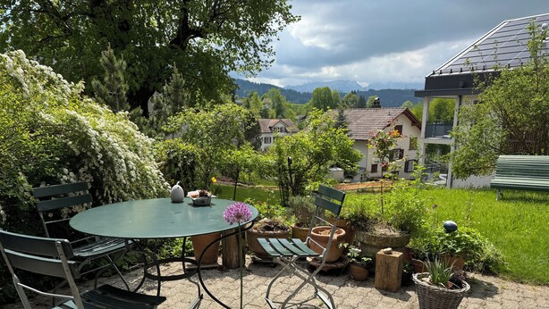 Une terrasse avec une table ronde et des chaises, entourée de plantes en pot et d’un banc.