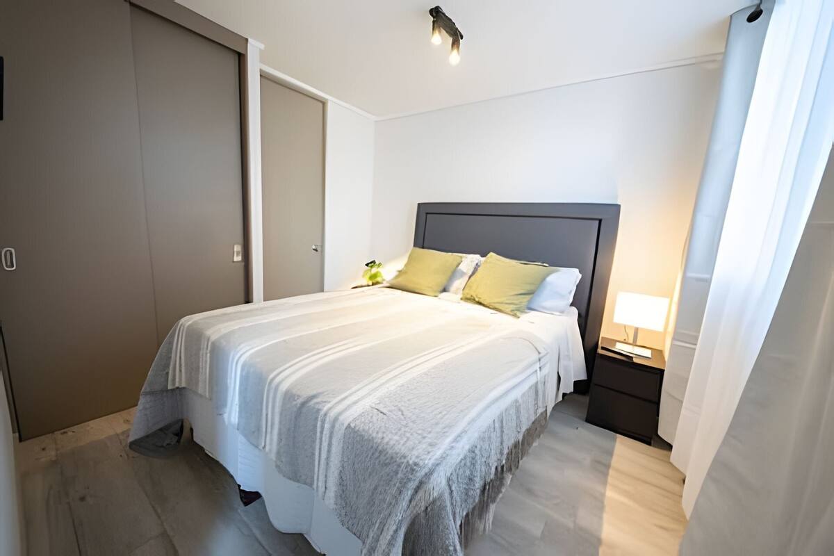 Ein ordentlich bezogenes Bett mit grauem Kopfteil, ein Nachttisch mit Lampe und eine Tür links.