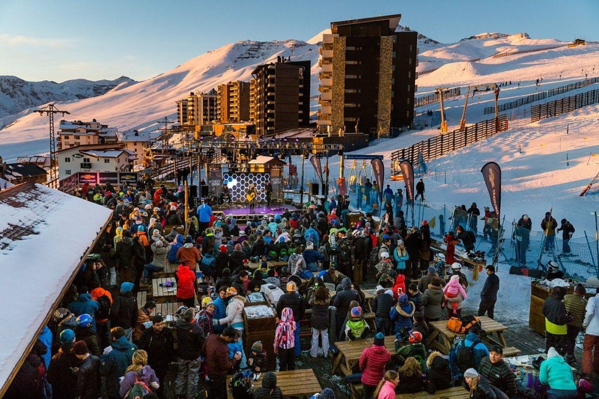 A crowded outdoor event with a stage and audience, set against a snowy mountain backdrop.