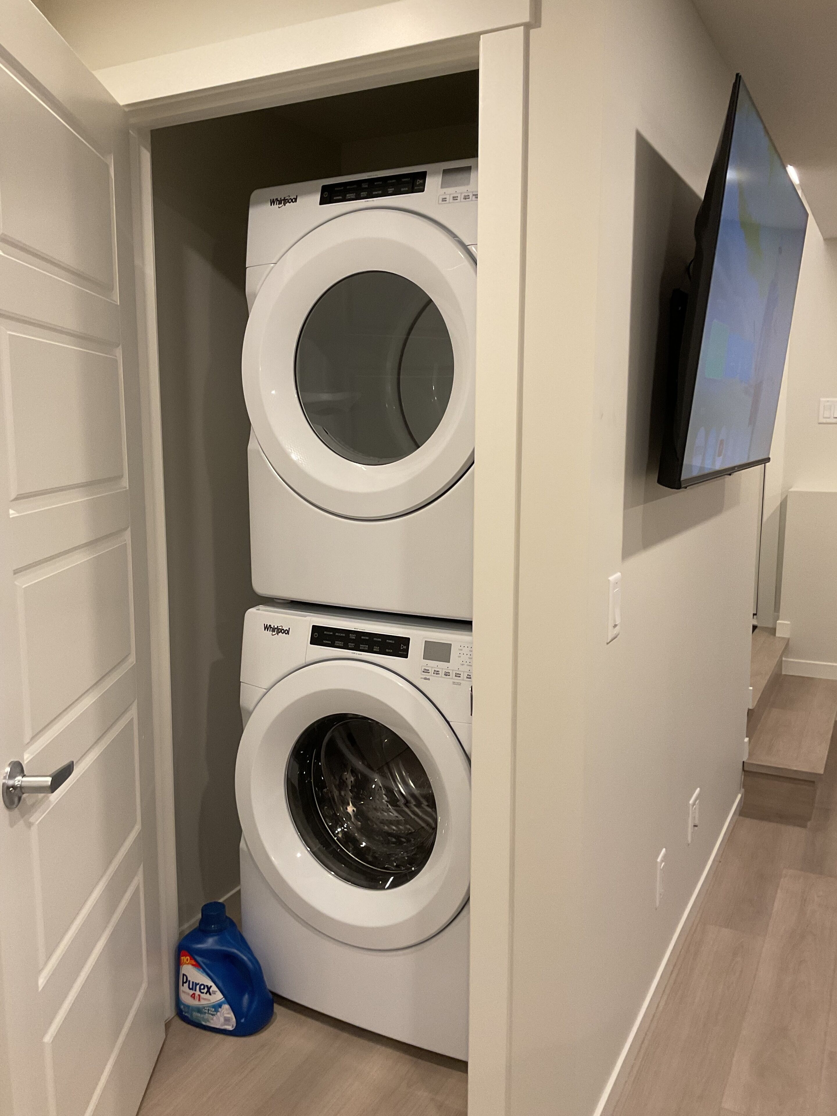 A compact laundry room with two front-loading washing machines, a television mounted on the wall, and a door leading to another room.