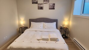 Una cama bien hecha con sábanas blancas y un cabecero gris, flanqueada por dos mesitas de noche con lámparas, en una habitación de diseño sencillo y limpio.
