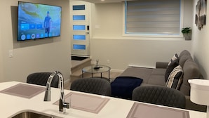 Una sala moderna con un televisor de pantalla plana montado en la pared, un sofá, una mesa de centro y un comedor con un fregadero de cocina.