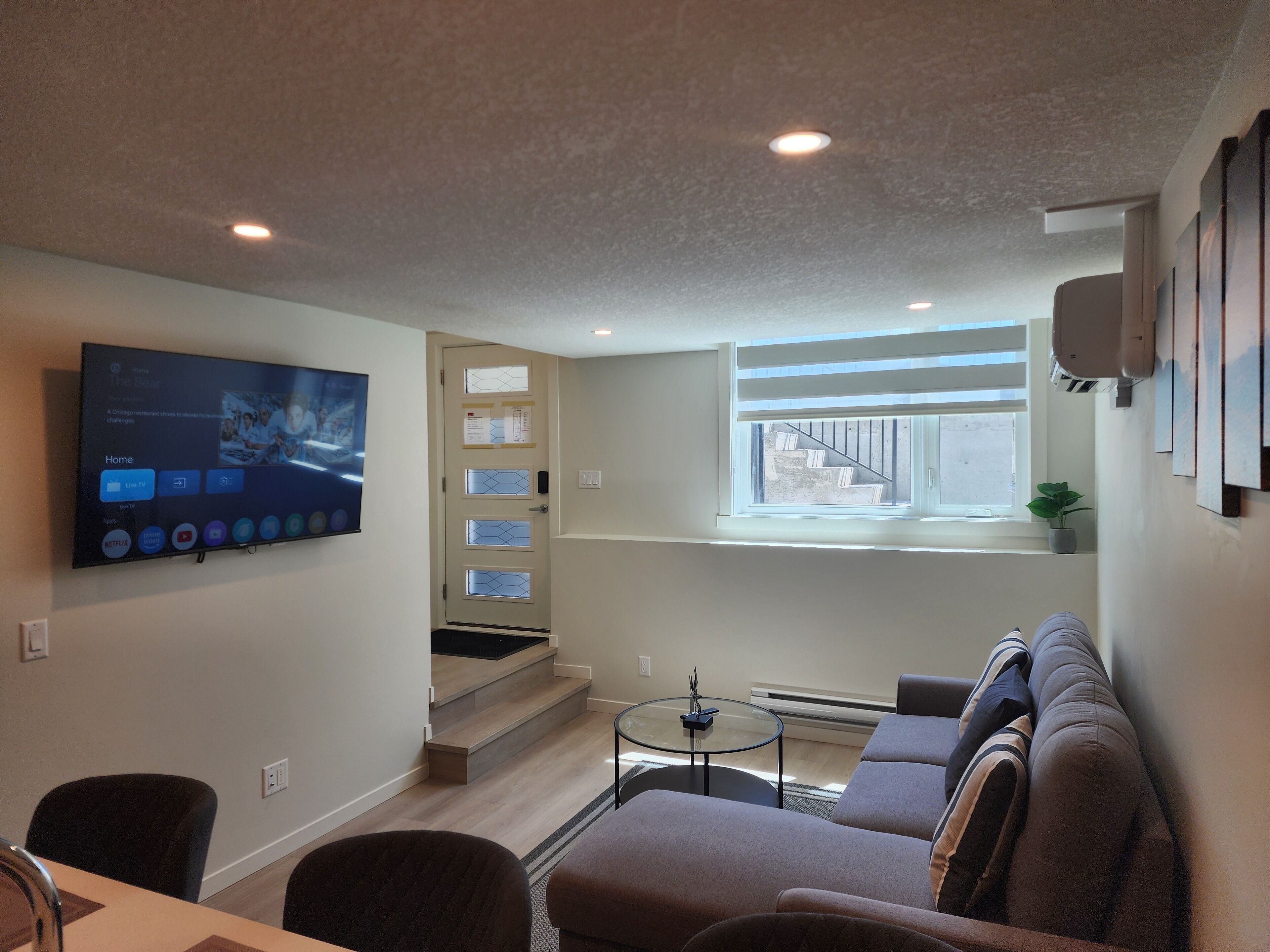 Una sala de estar moderna con un televisor de pantalla plana montado en la pared, un sofá, una mesa de centro y una escalera que lleva a un nivel superior.