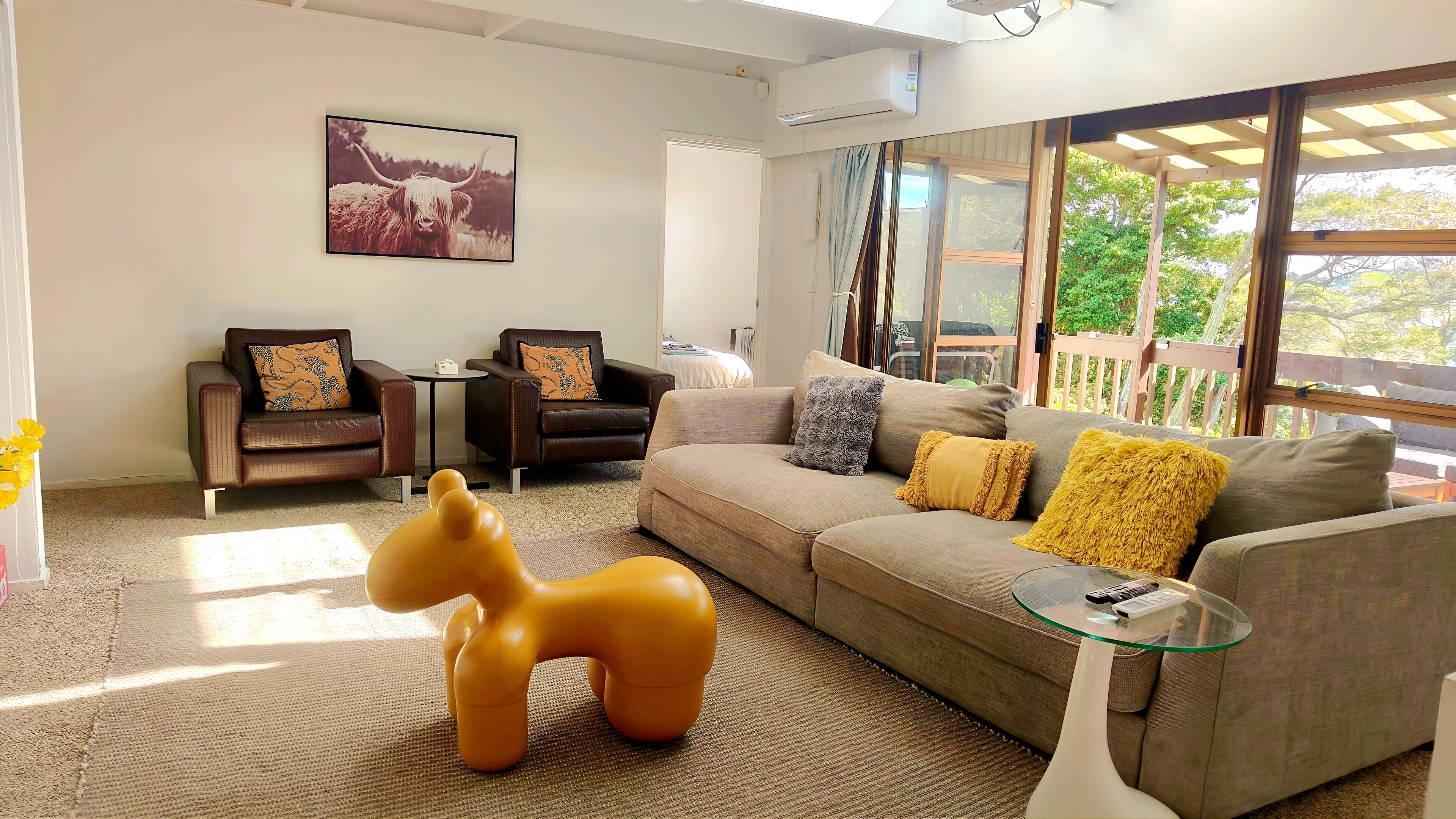 A living room with a sofa, two armchairs, a glass coffee table, a large window, and a mounted picture of a yak.