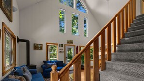 A spacious living area with a wooden staircase, large windows, and a cozy blue sofa. - Wildwood  Cabin in Welches (Mount Hood Village)