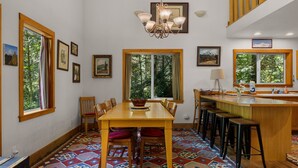 A dining area with a wooden table and chairs, a chandelier, and large windows with curtains. - Wildwood  Cabin in Welches (Mount Hood Village)