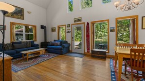 A spacious living room with a sofa, a coffee table, and a fireplace. There are large windows providing natural light and views of the outdoors. - Wildwood  Cabin in Welches (Mount Hood Village)