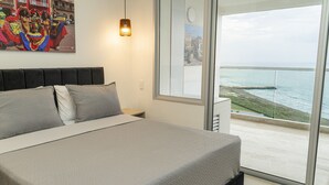 A bedroom with a large bed, a nightstand, a lamp, and a view of the ocean through a large window. - Loft Apartment 1510 (Cartagena de Indias)
