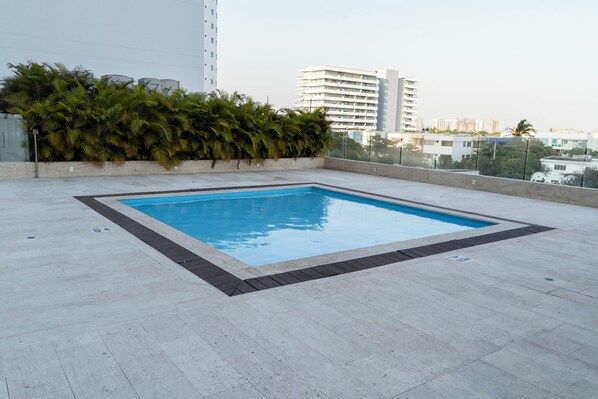 A rooftop pool with a clear blue pool, surrounded by a light-colored tiled deck and a low hedge.