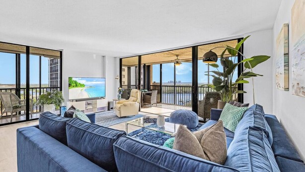 A spacious living room with a large sofa, a coffee table, and a TV. There is a balcony with a view of the sea.