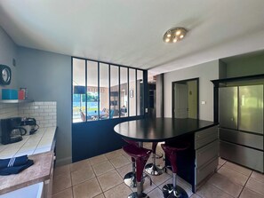 Eine moderne Küche mit einer zentralen Kochinsel, Barhocker, einem Kühlschrank und Blick durch ein großes Fenster.