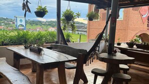 Un espacio comedor al aire libre cubierto con mesas y bancos de madera, plantas en macetas y vistas a un pueblo lejano y vegetación.