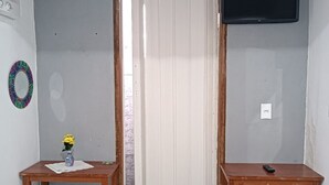 Una habitación con puerta de madera, un televisor fijado a la pared, dos mesitas de noche de madera, una pequeña planta en maceta y suelo de baldosas.