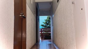 Un pasillo estrecho con una puerta de madera a la izquierda, suelo de baldosas y vistas al exterior con un portón rojo y vegetación.