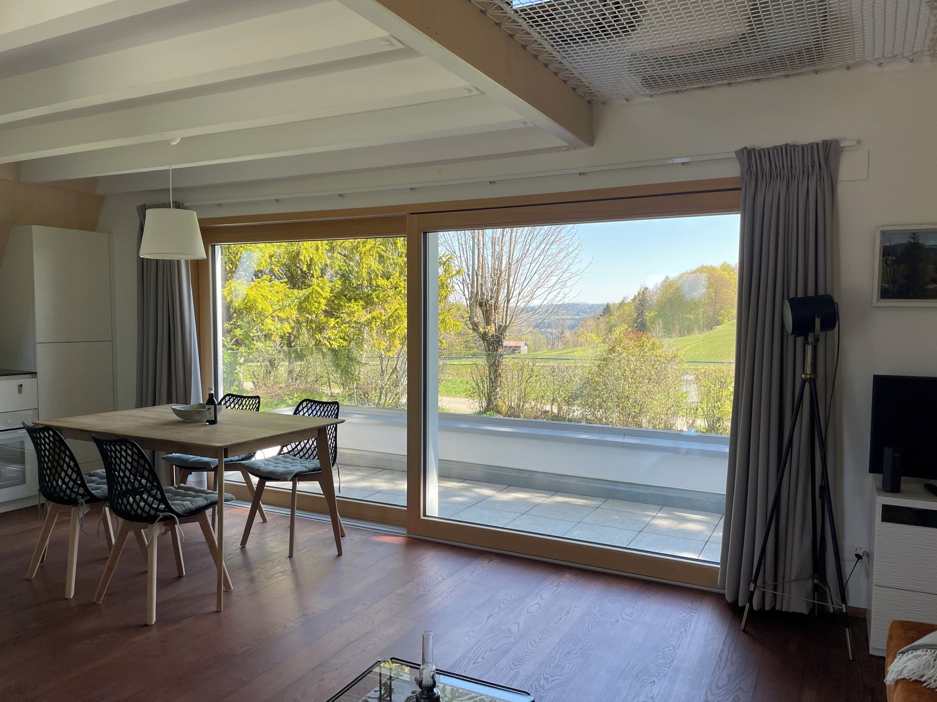 Een eetruimte met een tafel, stoelen, een lamp en uitzicht op een groen landschap door grote ramen.