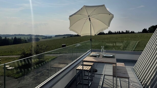 Ein Balkon mit Tisch für zwei Personen, einem großen Sonnenschirm und Blick auf eine grüne Landschaft.