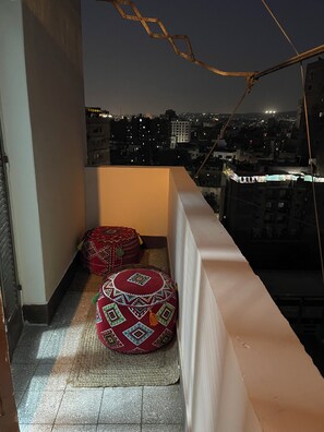 A balcony with two red patterned cushions, a view of a cityscape at night, and a railing.