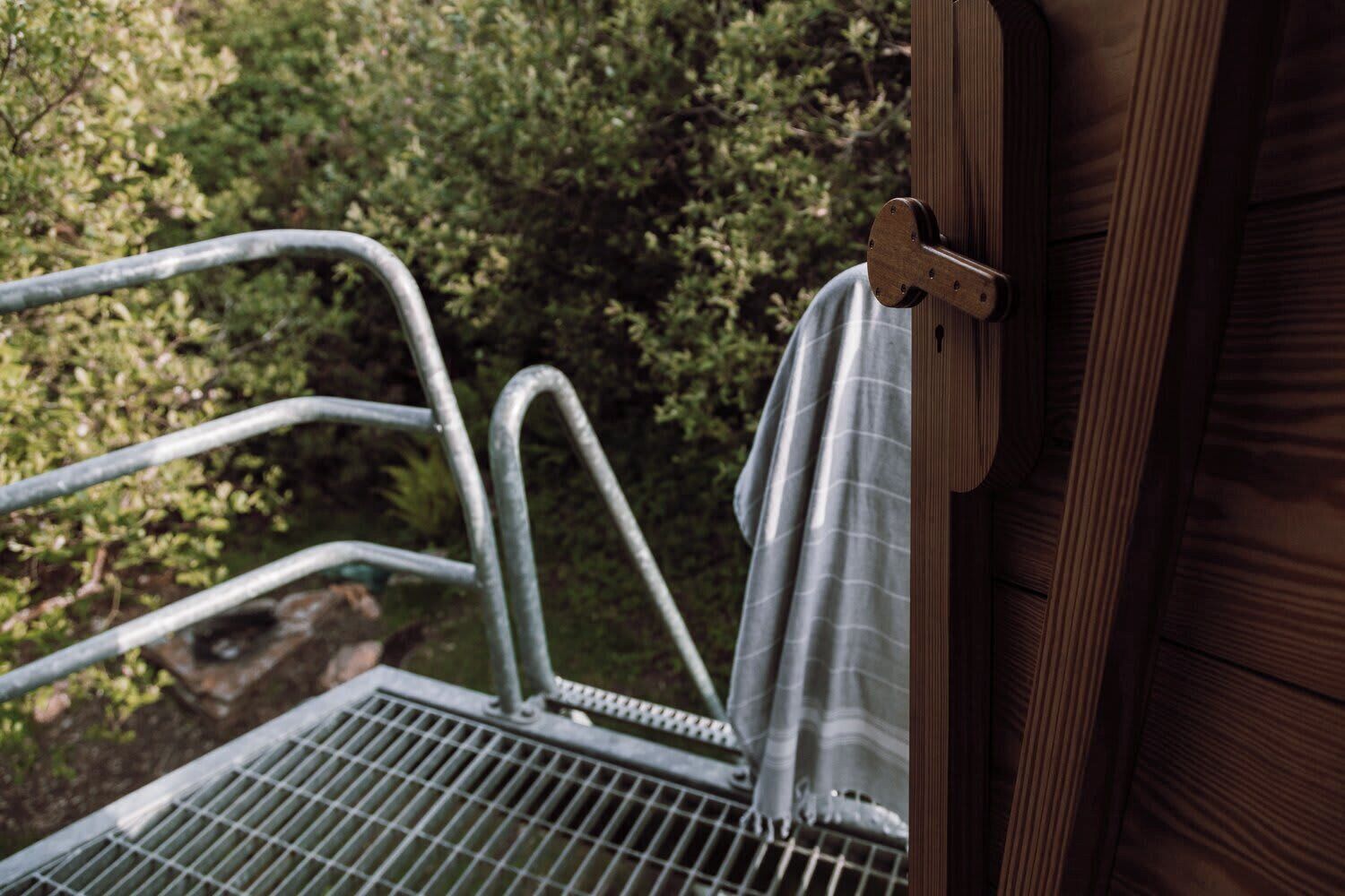 Un banc en métal installé à l’extérieur, doté d’une rambarde en métal, d’une porte en bois et offrant une vue sur la verdure en arrière-plan.