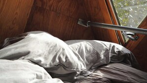 A bedroom with a slanted ceiling, a window with a metal handle, and a bed with a gray blanket.