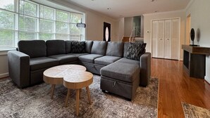 Un salon moderne avec un canapé d’angle, un pouf, une table basse ronde et une cheminée.