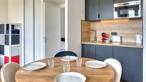 A compact kitchen with modern appliances, a dining area with a wooden table set for four, and a built-in shelving unit with storage. - Petit Castel - charmant T2 de 39m² au cœur de Rouen – Rue du Gros-Horloge ! (Rouen)