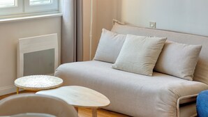 A beige sofa with two pillows, a small round table, and a radiator. - Petit Castel - charmant T2 de 39m² au cœur de Rouen – Rue du Gros-Horloge ! (Rouen)
