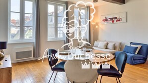 A modern living room with a dining area, large windows, and a blue sofa. - Petit Castel - charmant T2 de 39m² au cœur de Rouen – Rue du Gros-Horloge ! (Rouen)