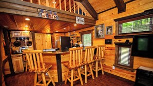Acogedor interior de cabaña con paredes y suelos de madera, una zona de bar con taburetes, una cocina con electrodomésticos y una chimenea.