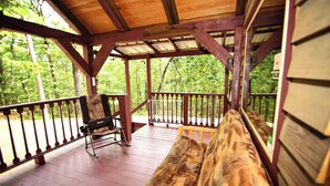 Una terraza cubierta de madera con bancos y una mecedora, rodeada por una barandilla de madera, con vistas a un bosque.