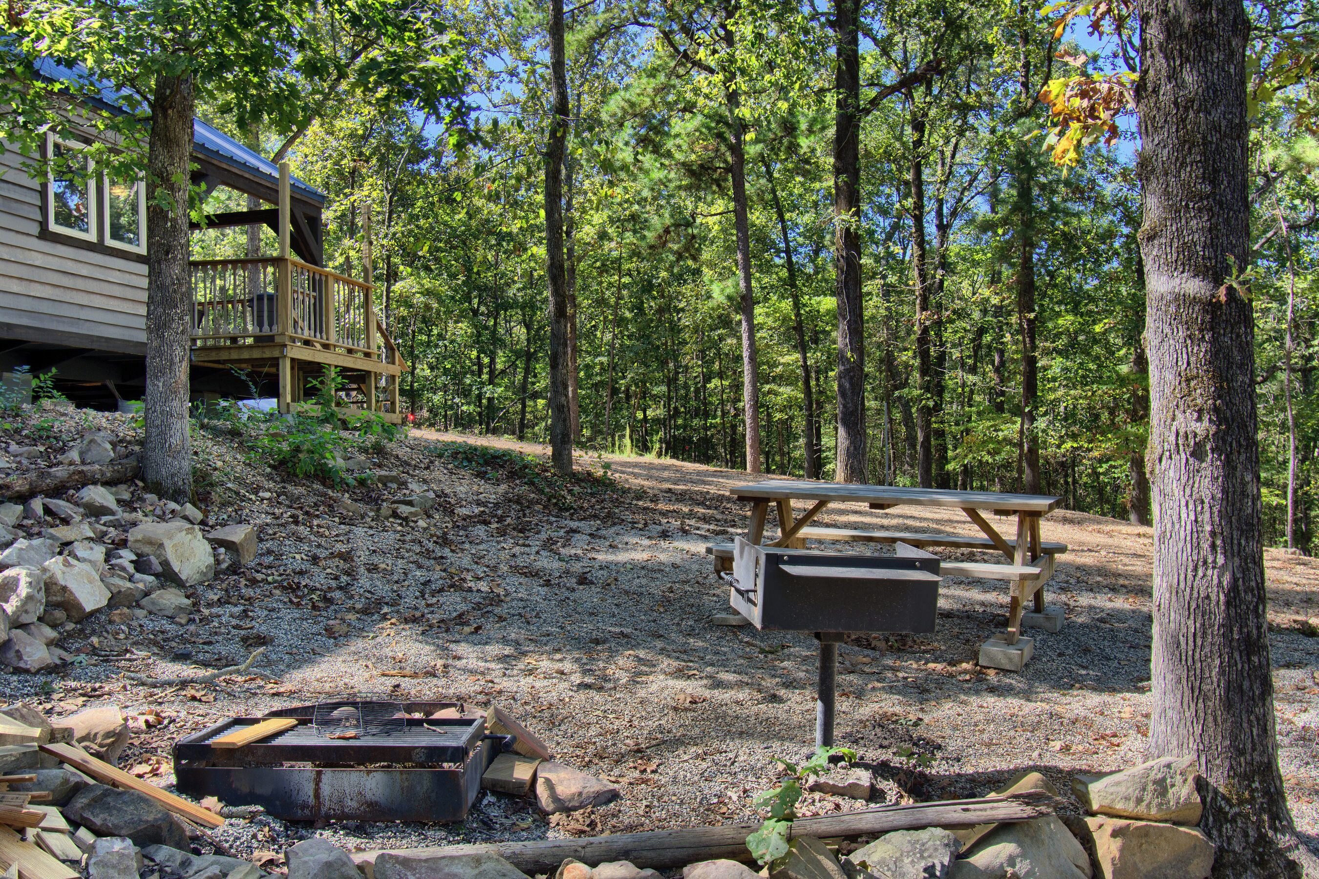 Une cabane avec une terrasse, une table de pique-nique et un foyer, située dans un cadre forestier.