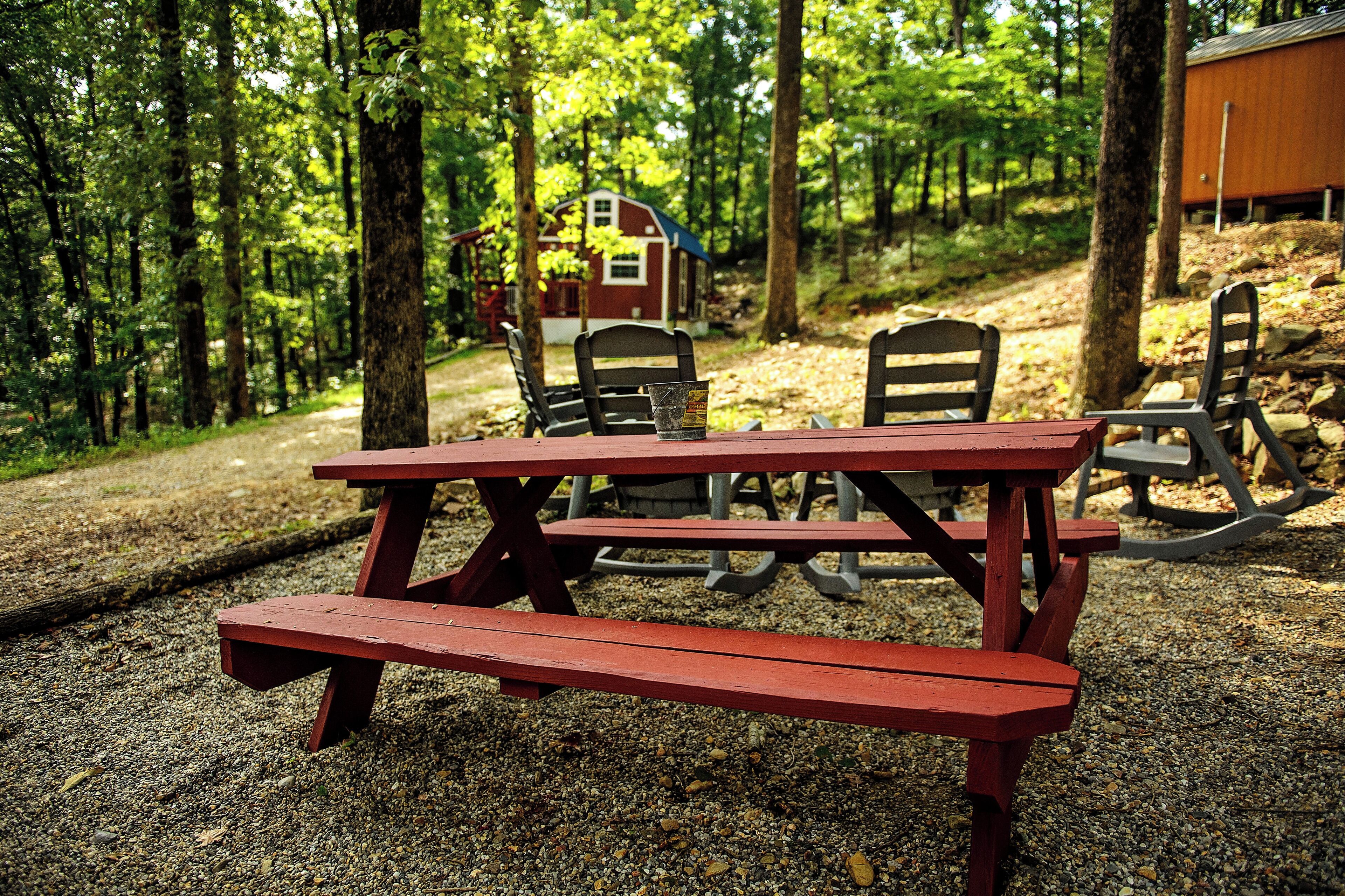 Una mesa de picnic con bancos en un área boscosa, con un edificio pequeño al fondo.