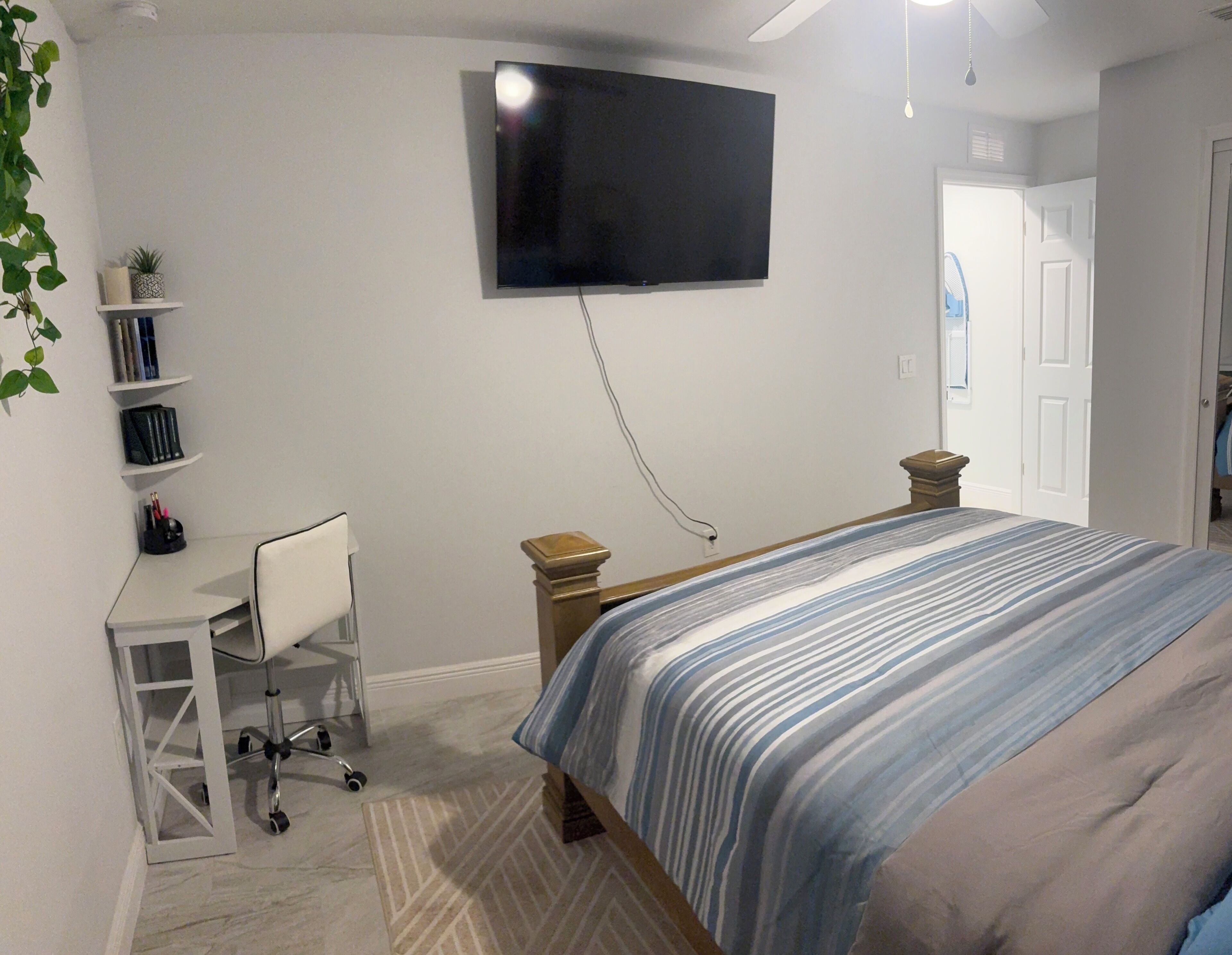 A bedroom with a bed, a desk, a chair, a TV mounted on the wall, and a door.