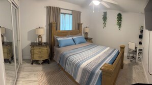 Un dormitorio con cama, mesitas de noche, ventilador de techo, televisión y ventana con cortinas.