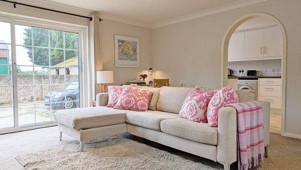 Een woonkamer met een beige hoekbank, een witte vloerkleed, een keuken met witte kasten en een groot raam met uitzicht op een auto en begroeiing.