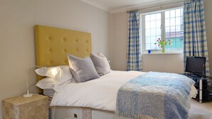 Een slaapkamer met een geel hoofdeinde, witte beddengoed, een bedlampje, een televisie en een raam met gordijnen.