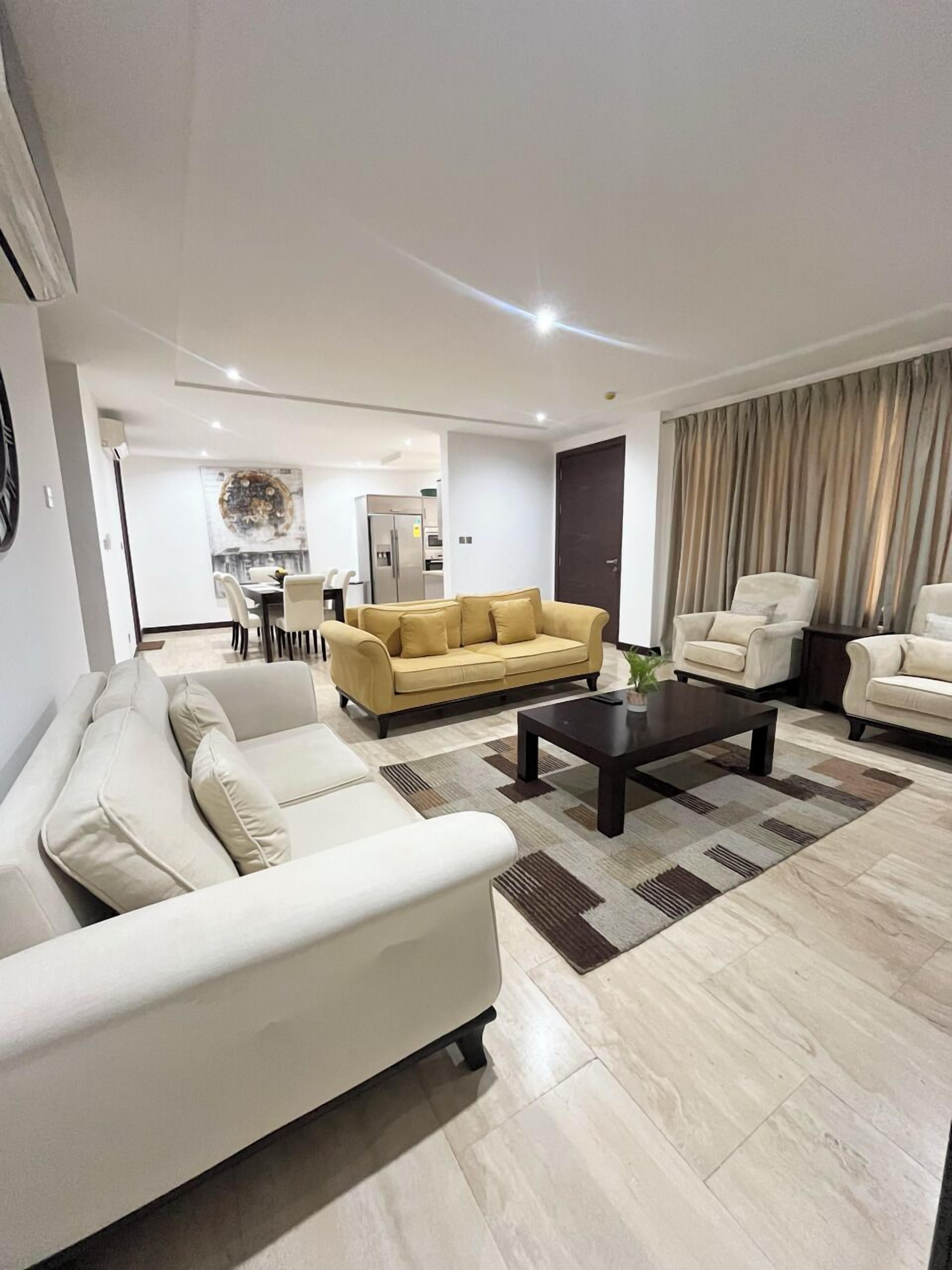 Ein modernes Wohnzimmer mit einer Couch, Sesseln und einem Couchtisch. Im Hintergrund ist ein Essbereich mit Tisch und Stühlen zu sehen.
