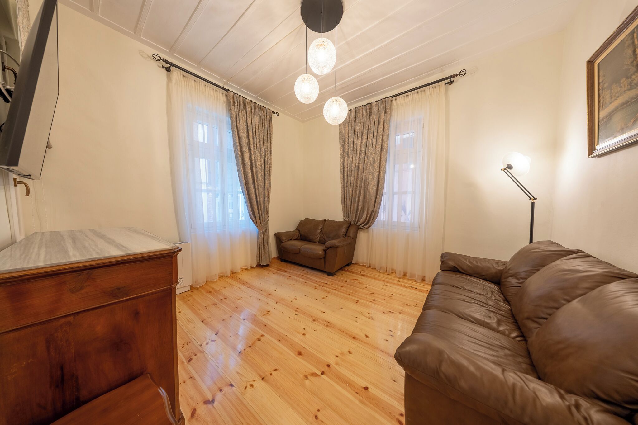 A room with a wooden floor, a brown leather sofa, a brown leather armchair, a wooden dresser, and a ceiling light fixture.