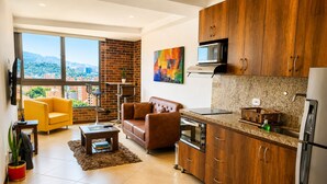 A modern living room with a sofa, armchair, and a coffee table. There is a kitchen area with a microwave, oven, and refrigerator. Large windows provide a view of the city.