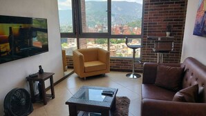 Una sala de estar con sofá, sillón y mesa de centro, ventanas amplias con vista a la ciudad y un televisor montado en la pared.