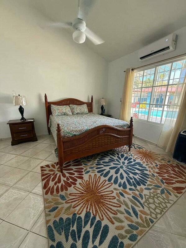 Un dormitorio con cama, ventilador de techo, unidad de aire acondicionado, ventana con cortinas y alfombra estampada.