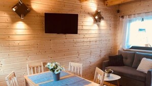 A cozy cabin interior with a wooden dining table, chairs, a sofa, a small side table, and a flat-screen TV mounted on the wall. - Cottage in Plesna Near Sandy Beach (gmina Będzino)
