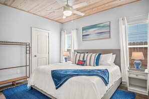 Une chambre à coucher avec un lit, un ventilateur de plafond, une fenêtre avec des stores, une porte et un tableau au mur.